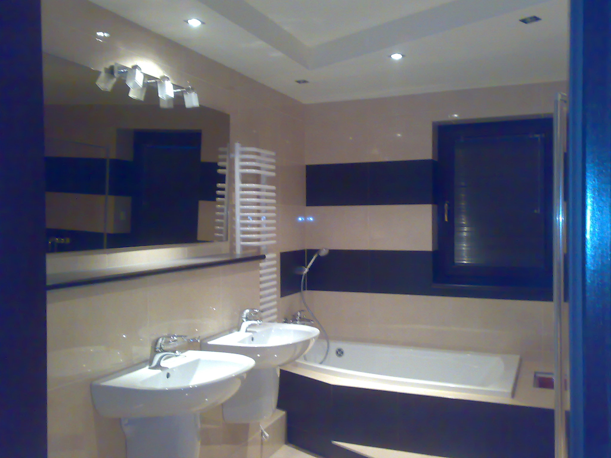 Ukończona łazienka z dwoma umywalkami na postumentach, białą wanną obudowaną ciemną płytą i grzejnikiem drabinkowym na ścianie w beżowo-czarne pasy, oświetlenie halogenowe sufitowe i nad lustrem.